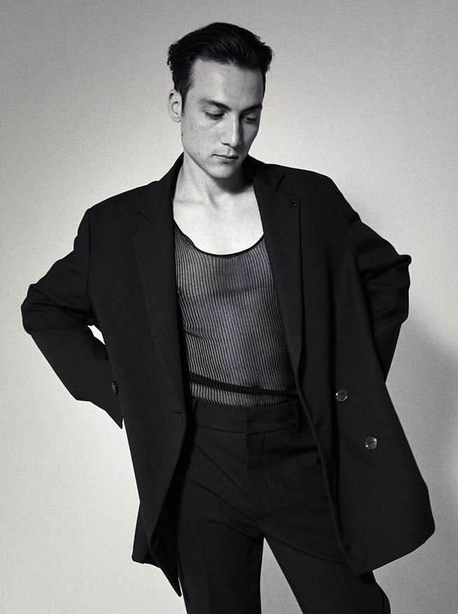 A stylish portrait of an adult male model in a black suit and sheer top, posing in a photo studio with soft, neutral lighting.