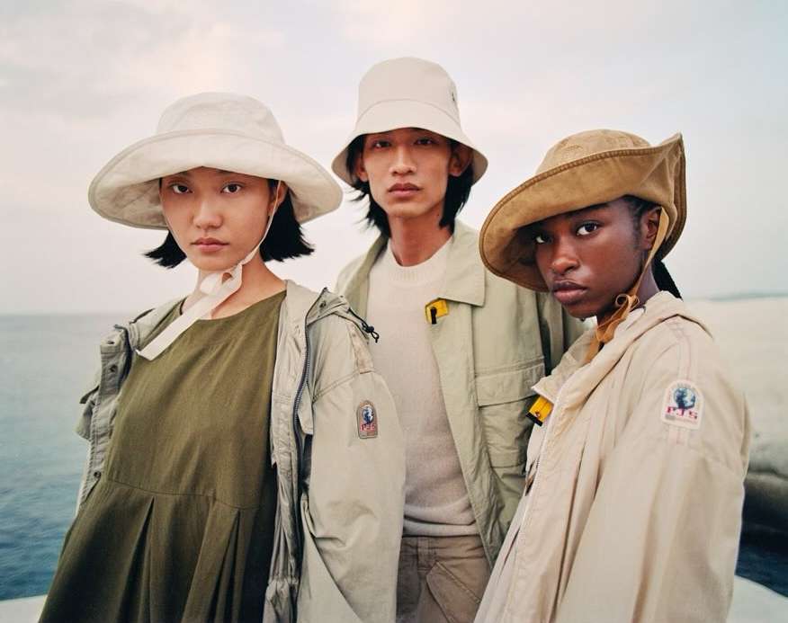 Three diverse fashion models wearing hats and jackets pose outdoors in front of water and sky with neutral color grading.