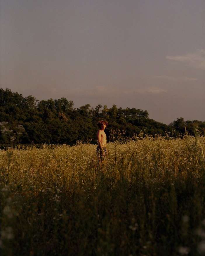 Person standing in a field of flowers. There is a line of trees in the background, and the sky is overcast and gray. The person is small in frame.