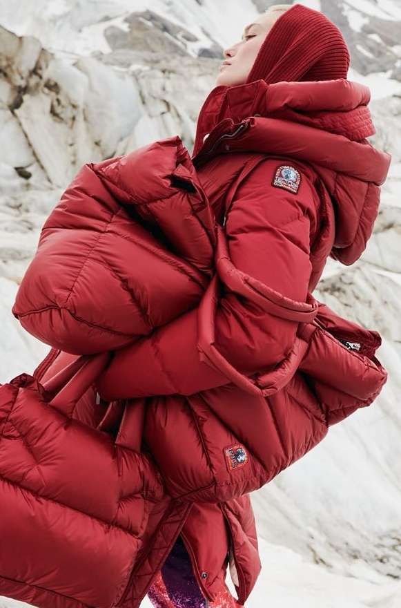Fashion shot of a woman in a red puffer jacket with a hood, posing outdoors against a snowy, mountainous landscape in soft lighting.