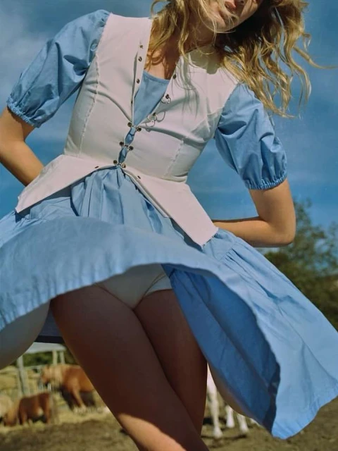 Young woman in a light blue dress and white vest, stands outdoors with horses in the background on a sunny day.