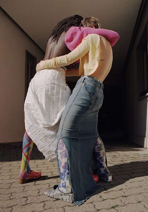 Two women embrace in a sunlit outdoor setting, showcasing colorful tights, denim, and white clothing, highlighting unique style and textures.