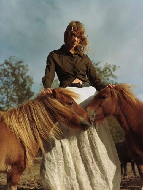 Woman in a brown shirt and white skirt standing outdoors with two brown ponies in a field under a cloudy sky in a portrait shot.