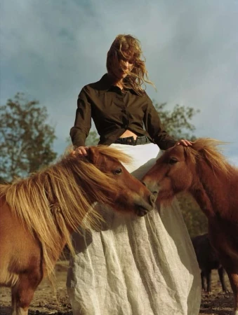 Woman in a brown shirt and white skirt standing outdoors with two brown ponies in a field under a cloudy sky in a portrait shot.