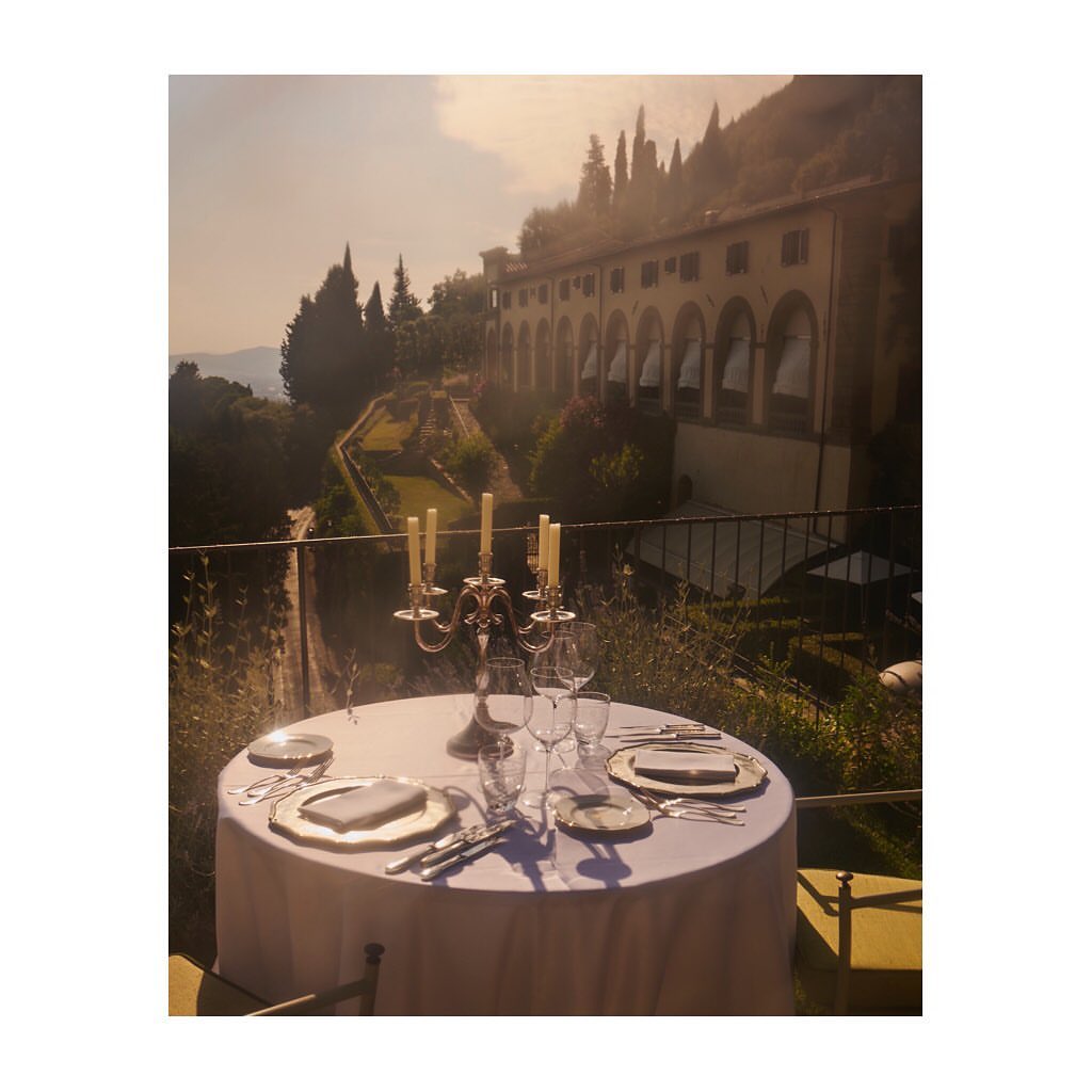Outdoor dining table set with silverware, glassware, and candelabra, set against a background of a grand building and lush greenery.