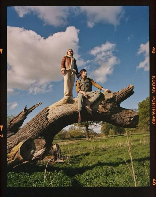 Two adult humans pose on a fallen tree trunk in a meadow under a cloudy sky. One stands wearing a brown jacket, the other sits. 