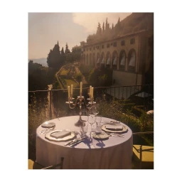 A set dining table with a candelabra on an outdoor terrace overlooking a grand estate building and hills.