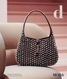 A dark crystal-embellished leather handbag resting on a light beige upholstered chair with a soft pink background.