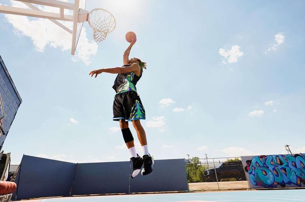 A man in sports gear leaps with a basketball near a hoop, set against a sunny sky and graffiti-covered wall in an urban outdoor setting.