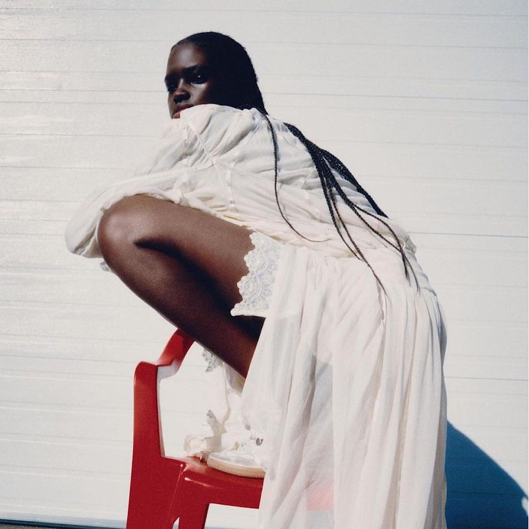 Portrait of a dark-skinned woman in a long white dress, sitting on a red chair, with a white garage door as the backdrop.