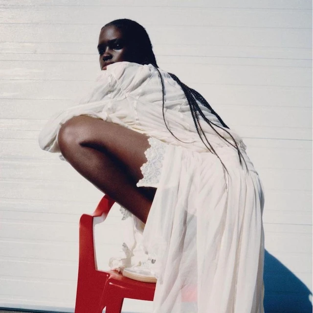 Portrait of a dark-skinned woman in a long white dress, sitting on a red chair, with a white garage door as the backdrop.