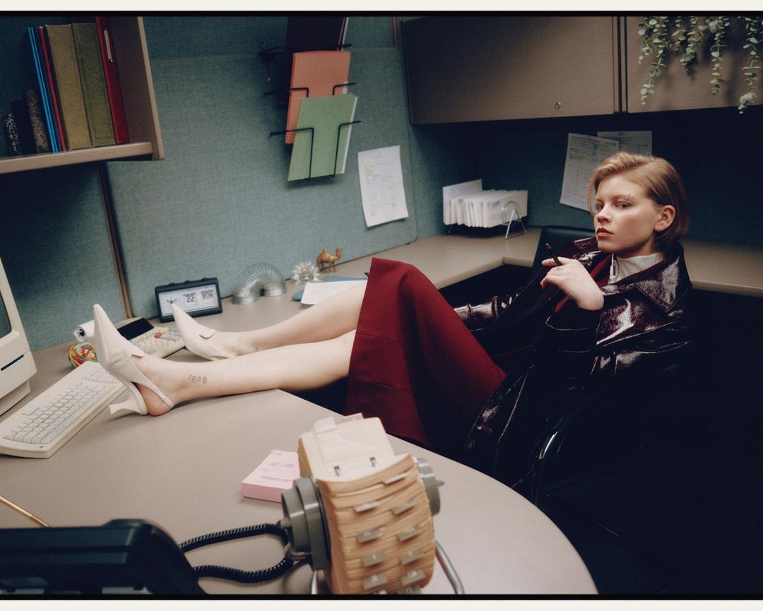 Stylish woman poses in an office cubicle wearing patent leather coat, red skirt, white heels, with an obsolete computer and rotary phone.