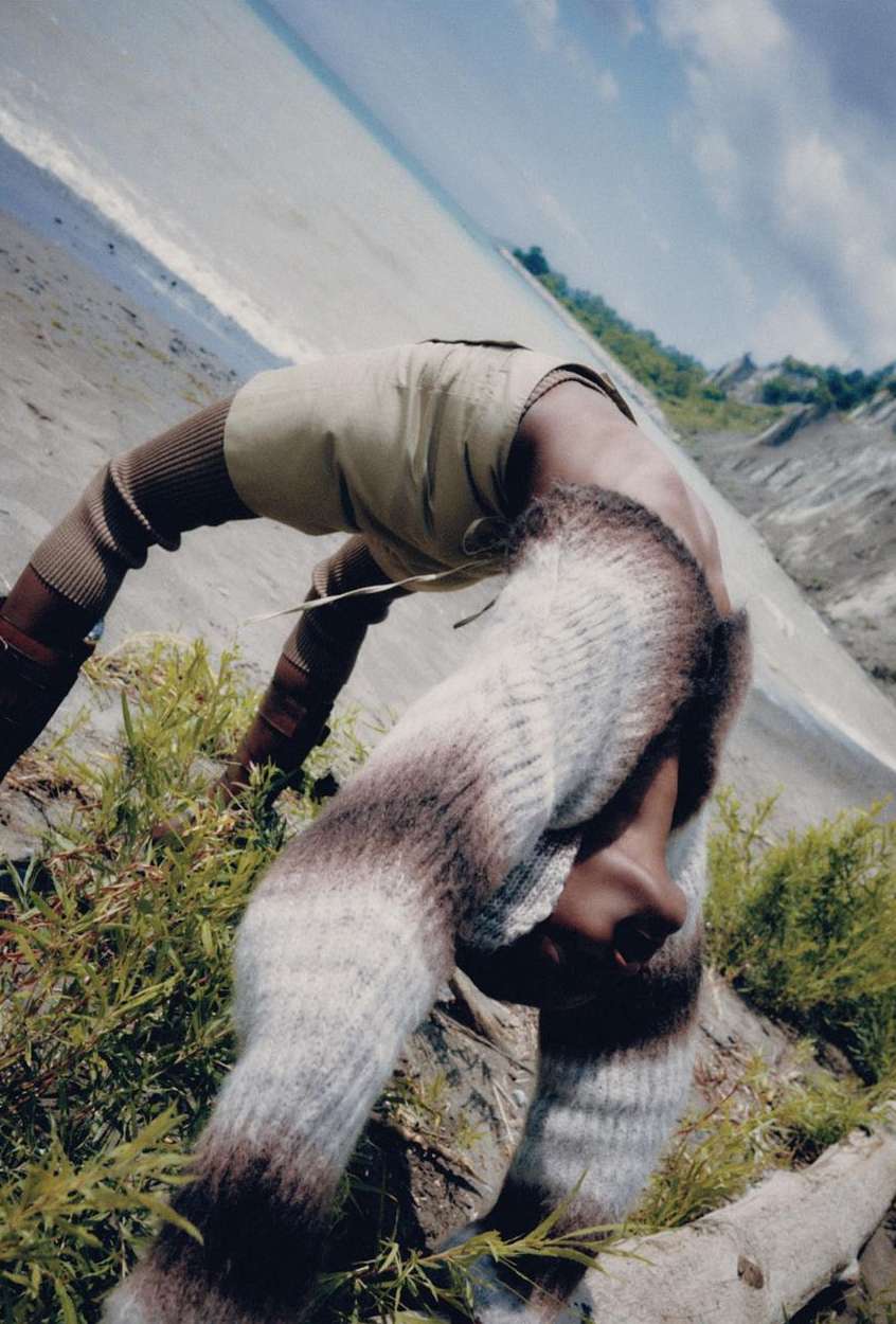 Model in a backbend pose outdoors on a rocky beach, wearing knit leggings, with the horizon line angled sharply in the background.