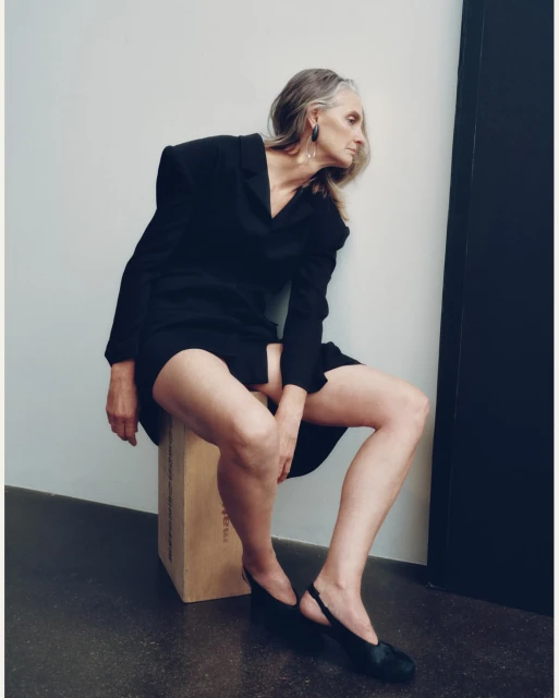 Elegant older woman in black blazer and skirt posing indoors, seated on a box with black slingback heels, looking off-camera.
