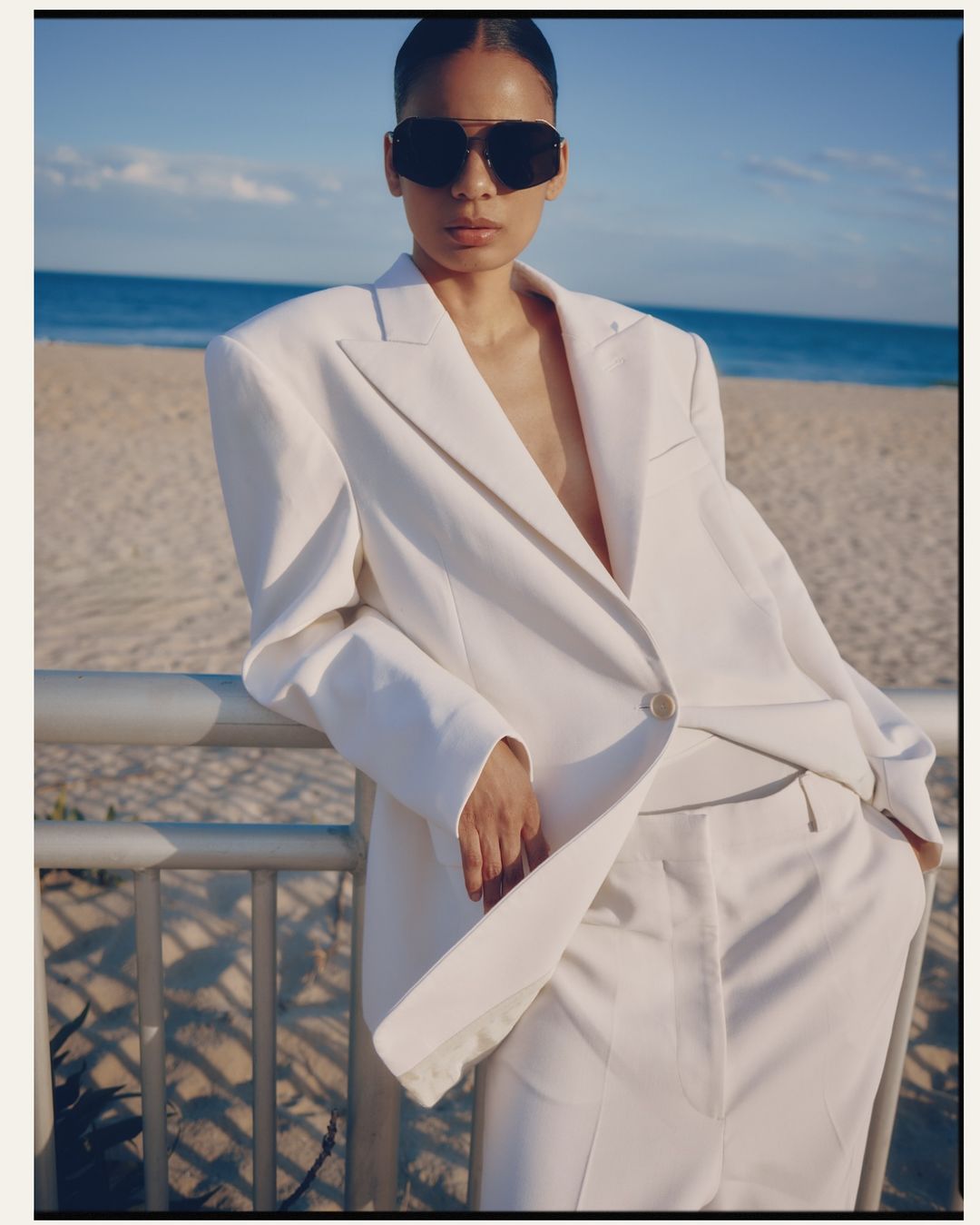 A fashionable adult female in a white suit and sunglasses poses on a beach with ocean and sky background during daytime in a medium shot.
