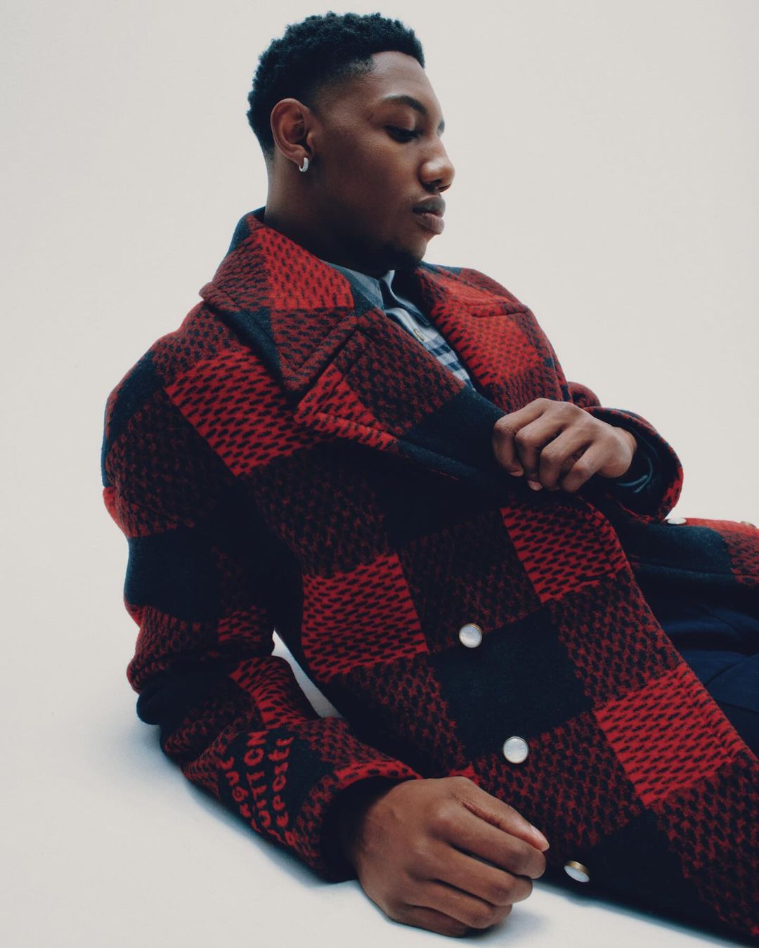 Young man with dark hair and a red and black checkered coat poses indoors against a white background, looking down and to the right.