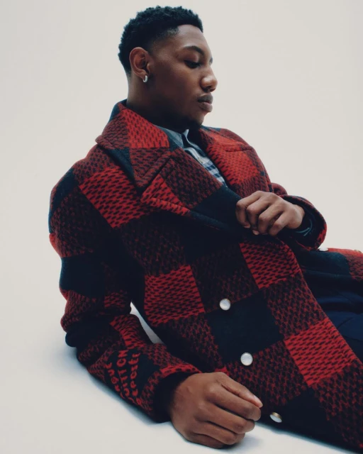 Young man with dark hair and a red and black checkered coat poses indoors against a white background, looking down and to the right.
