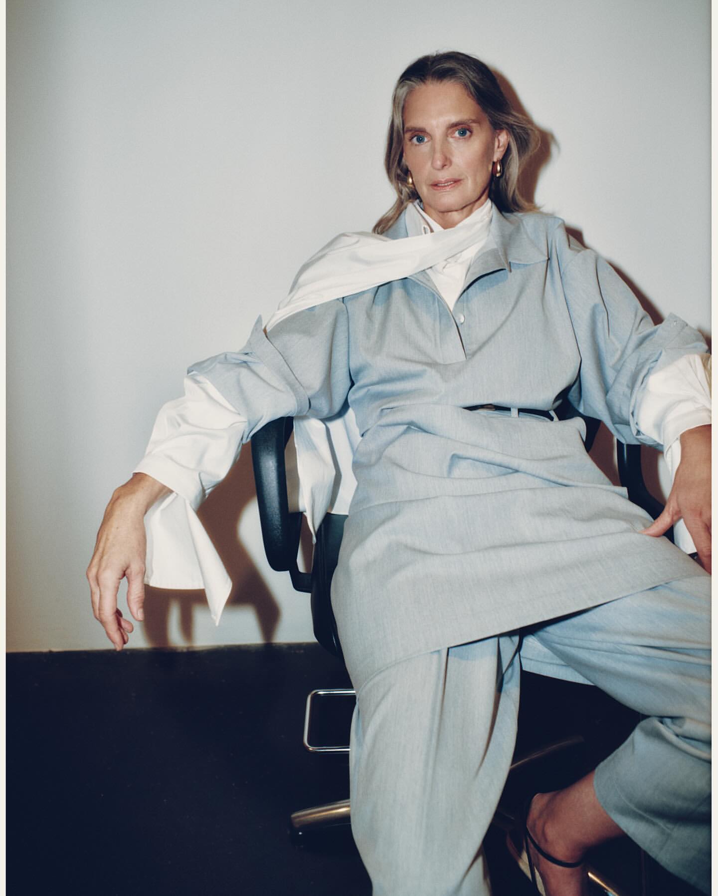 Older woman in a light blue pant suit sitting on a chair in a studio setting, posing for fashion portrait. Neutral wall background.