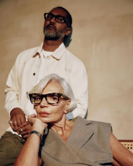 Stylish portrait of an adult man and an older woman wearing glasses and fashionable clothing in a brightly lit indoor setting.
