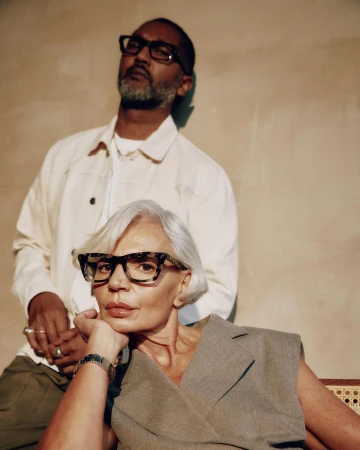 Stylish portrait of an adult man and an older woman wearing glasses and fashionable clothing in a brightly lit indoor setting.