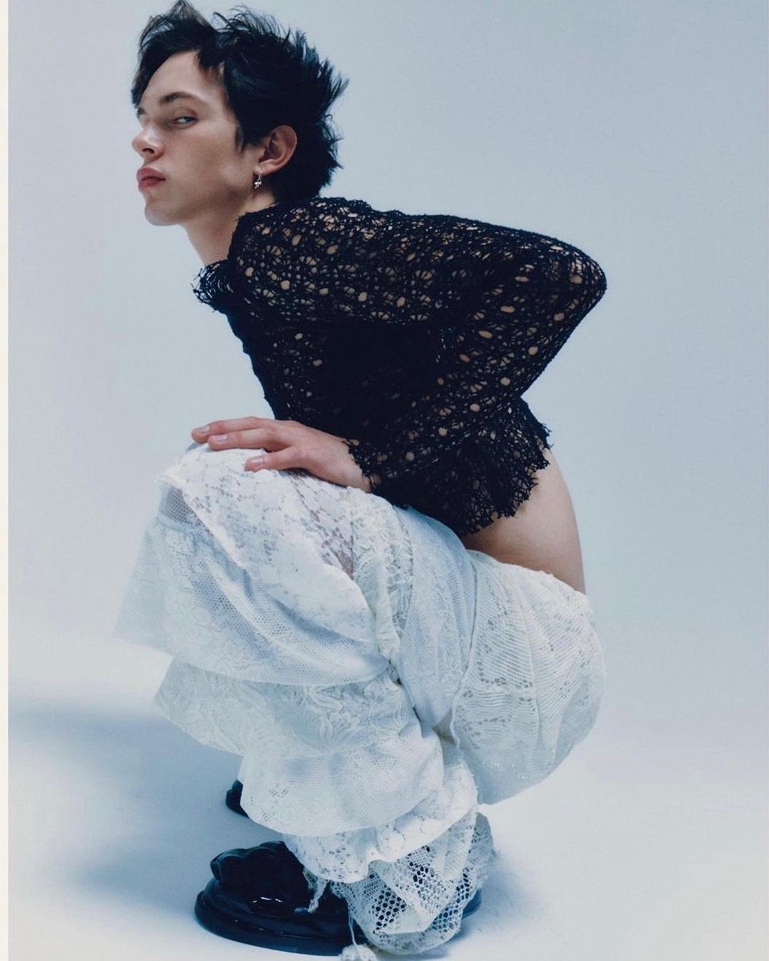 Young adult crouches wearing a black lace top and white lace pants in a studio with bright lighting.