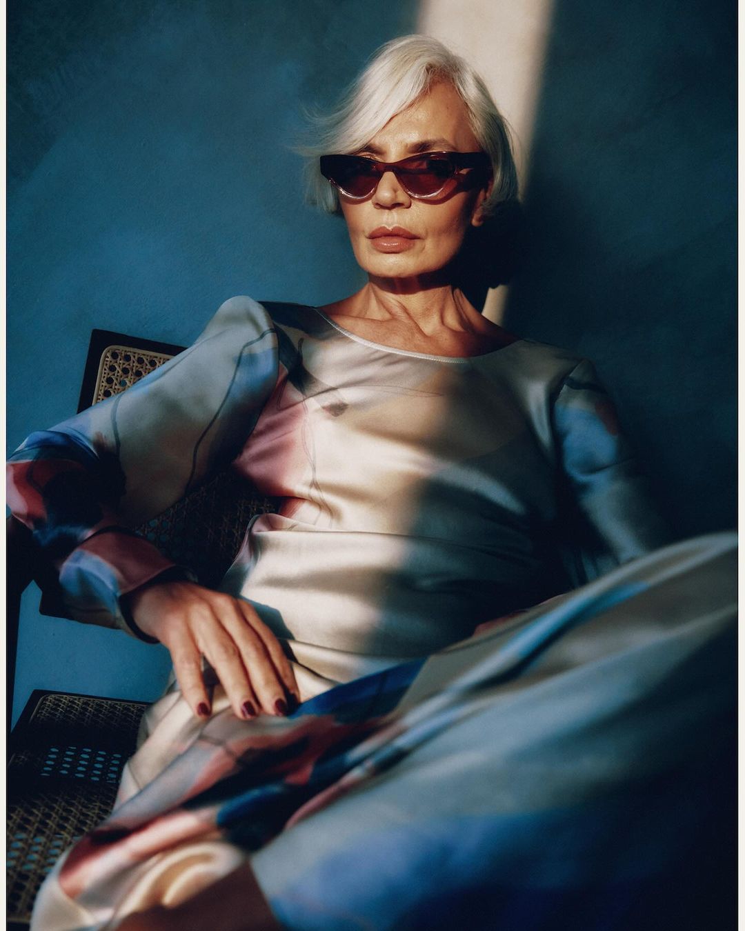 Elegant mature woman with short grey hair wearing stylish sunglasses and a patterned shirt, sitting in a chair against a blue wall.