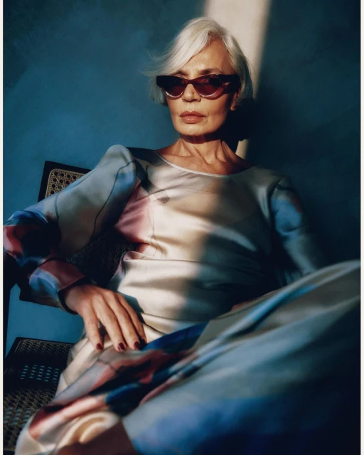 Elegant mature woman with short grey hair wearing stylish sunglasses and a patterned shirt, sitting in a chair against a blue wall.