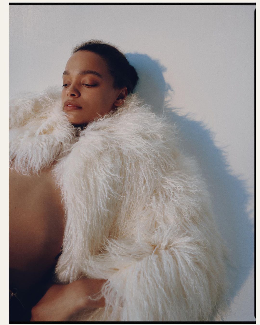 Woman poses in a shaggy off-white coat in an eye-level shot against a neutral background. Soft studio lighting enhances the textured garment.