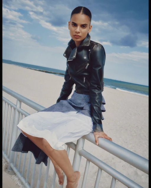 A woman in a black leather jacket and layered skirt sits on a railing at the beach on a sunny day with a cloudy sky.