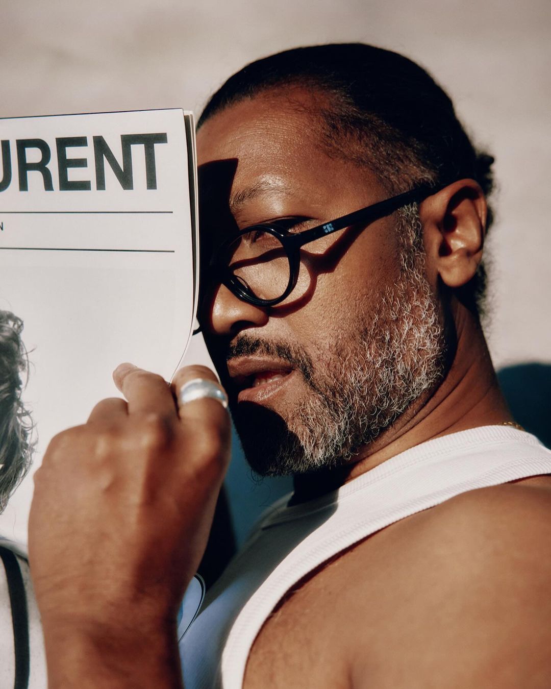 Man holding a magazine in front of his face wearing black glasses and a white tank top by Garrett Naccarato