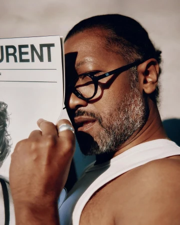 Man holding a magazine in front of his face wearing black glasses and a white tank top by Garrett Naccarato