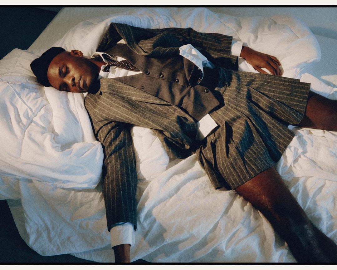 A young Black man sleeps in a pinstripe suit and pleated skirt on a white bed with a white collared shirt and dotted tie visible.