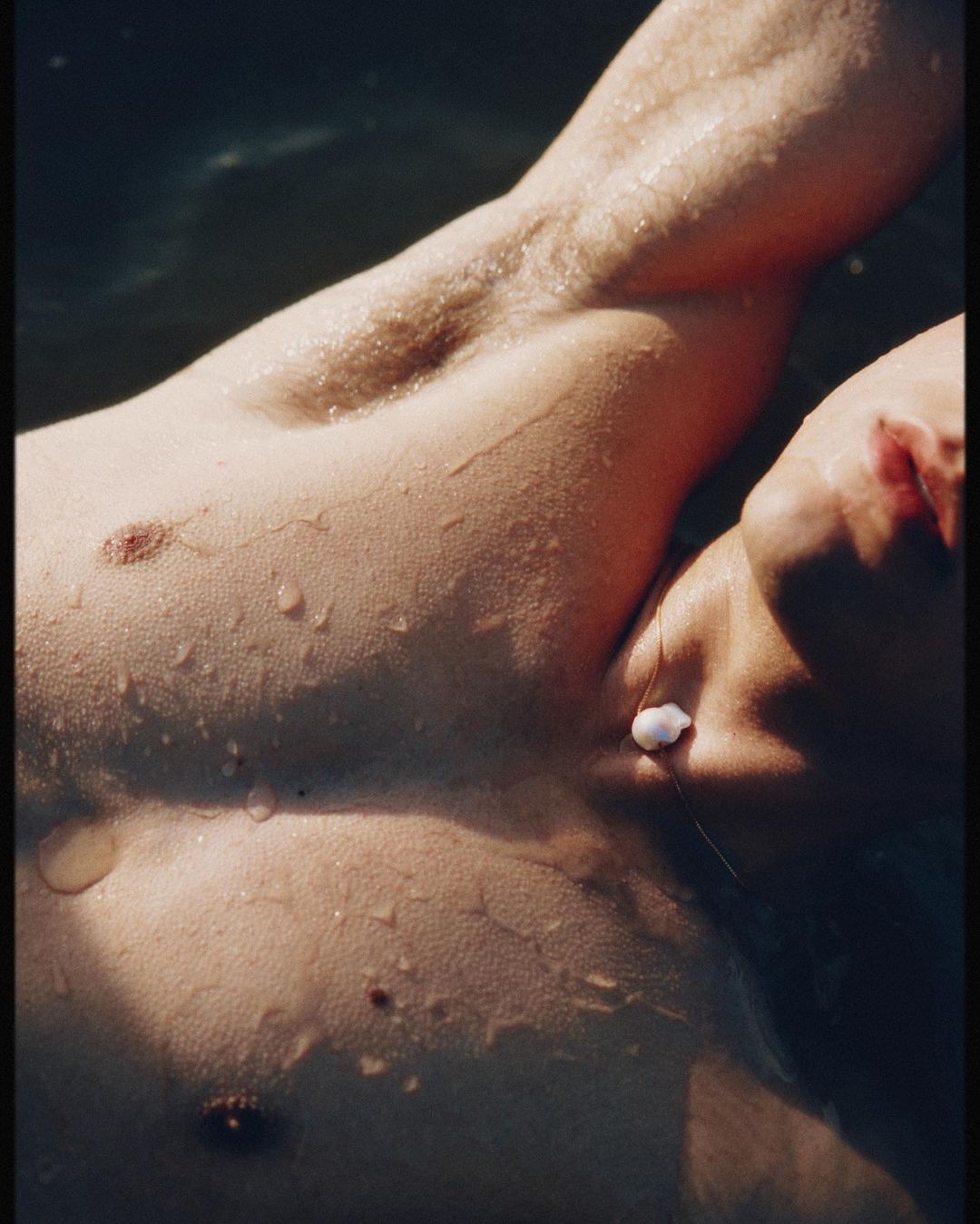 Close-up shot of a male torso in water with droplets of water on his skin. A small seashell sits on the man's chest.