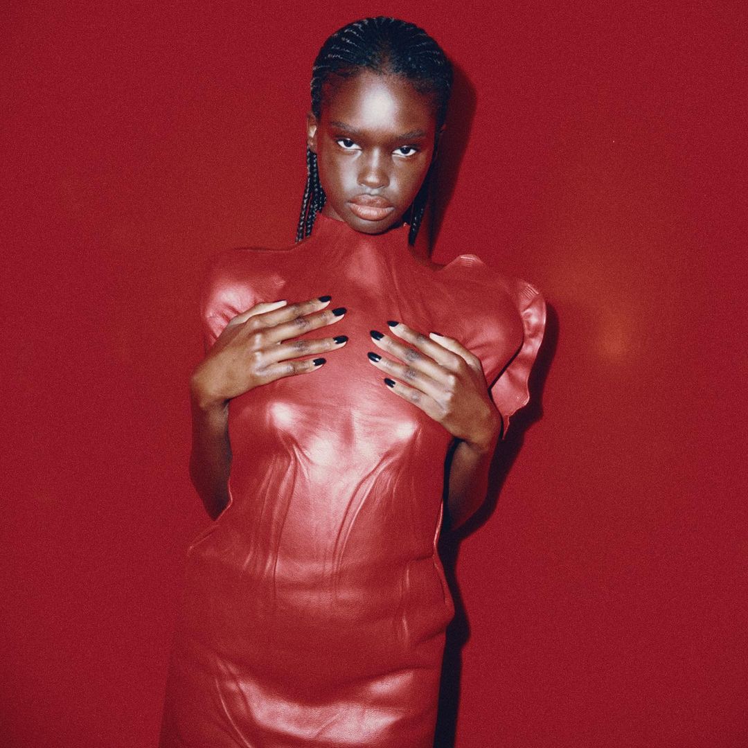 An African-descent female fashion model wearing a red sculpted dress poses in front of a matching seamless background in studio lighting.