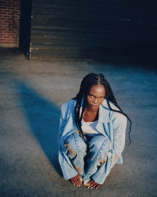 Young woman with long braided hair, wearing a light blue denim jacket and jeans, poses in an urban setting with a brick wall backdrop.