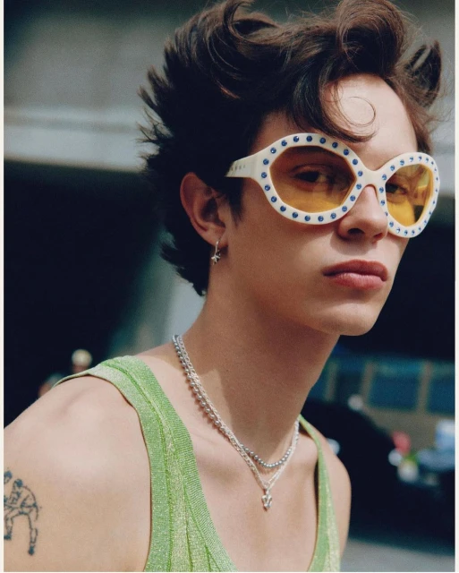 Close-up of a young adult male wearing sunglasses and a green top, posing in an outdoor setting with a blurred background.