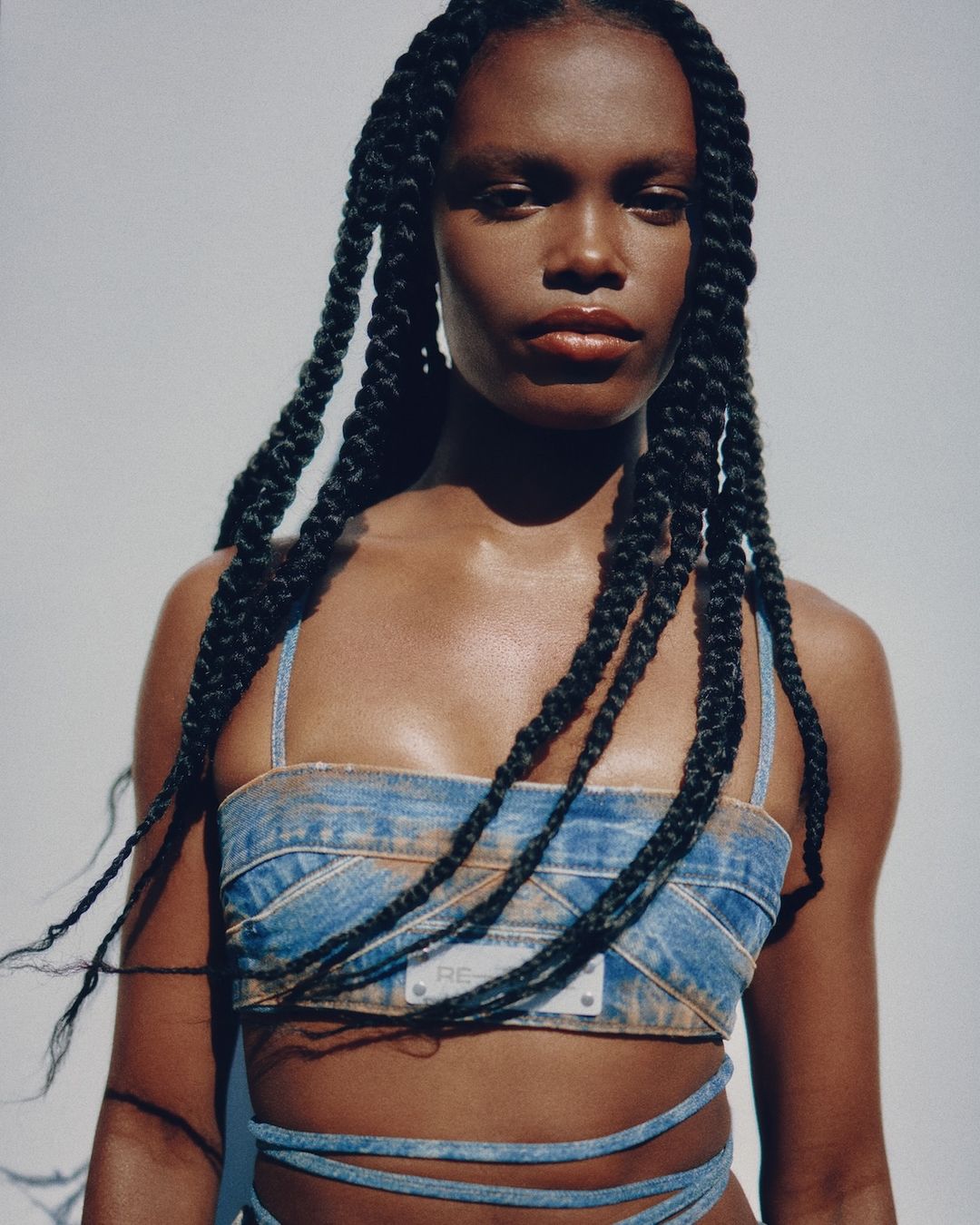 Fashion portrait of a woman with long braided hair wearing a denim top with a serious facial expression against a white background.