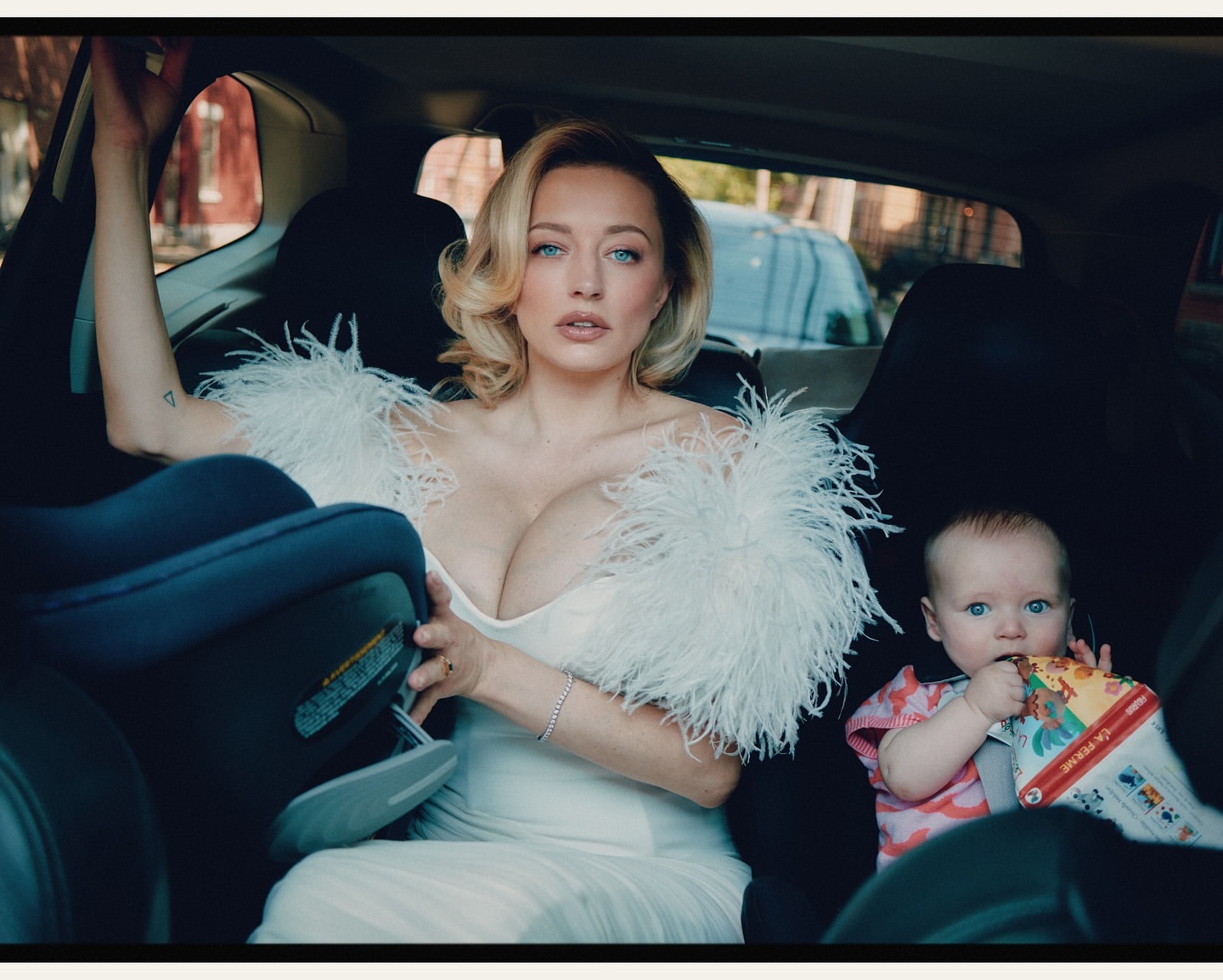 Blonde woman and child in car. Woman wears white dress with feather trim. Child eats with colorful book visible, seated in car seat.