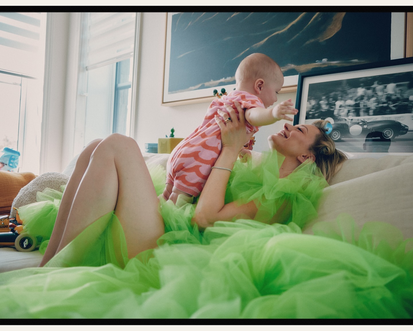 Blond woman in a neon green dress holds a baby in a pink patterned outfit while reclining on a cream-colored couch in a bright living room.