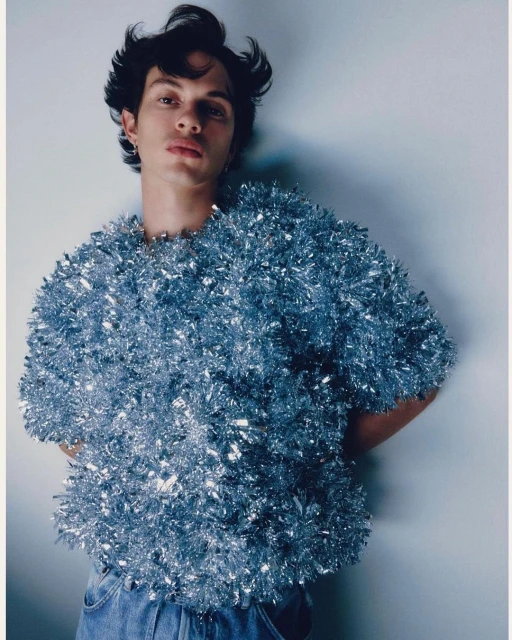 Portrait of a young adult male wearing a shiny, textured tinsel top and jeans against a pale background in a studio setting.