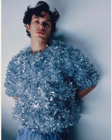 Portrait of a young adult male wearing a shiny, textured tinsel top and jeans against a pale background in a studio setting.