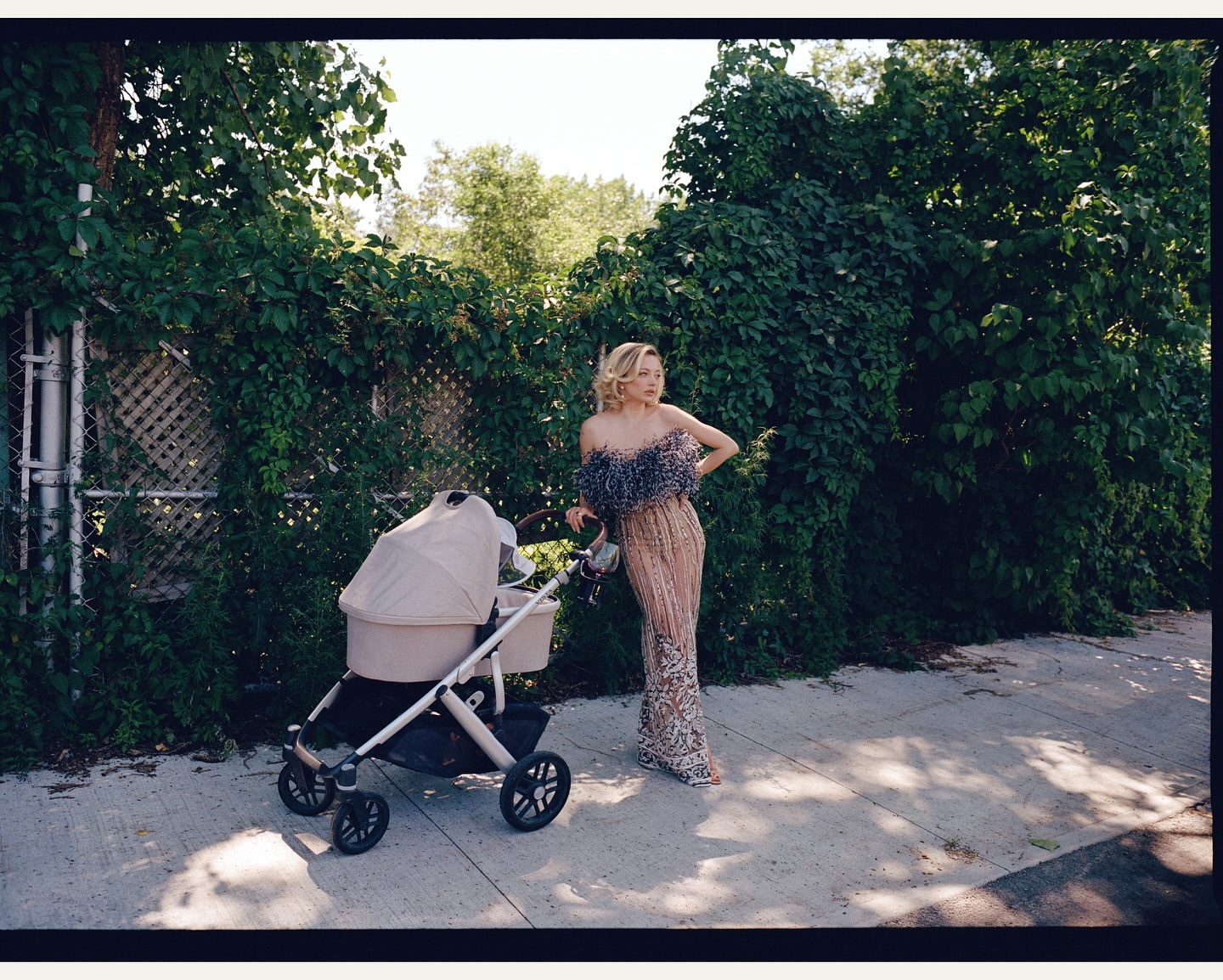 A woman in an elegant gown poses with a stroller on a city sidewalk, framed by lush greenery and a sunny ambiance, creating a striking contrast.