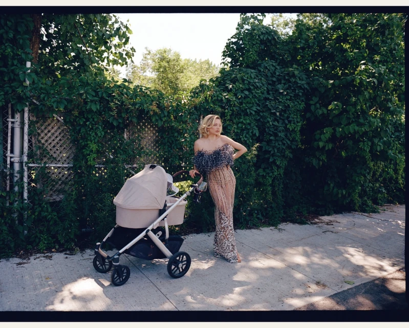A woman in an elegant gown poses with a stroller on a city sidewalk, framed by lush greenery and a sunny ambiance, creating a striking contrast.