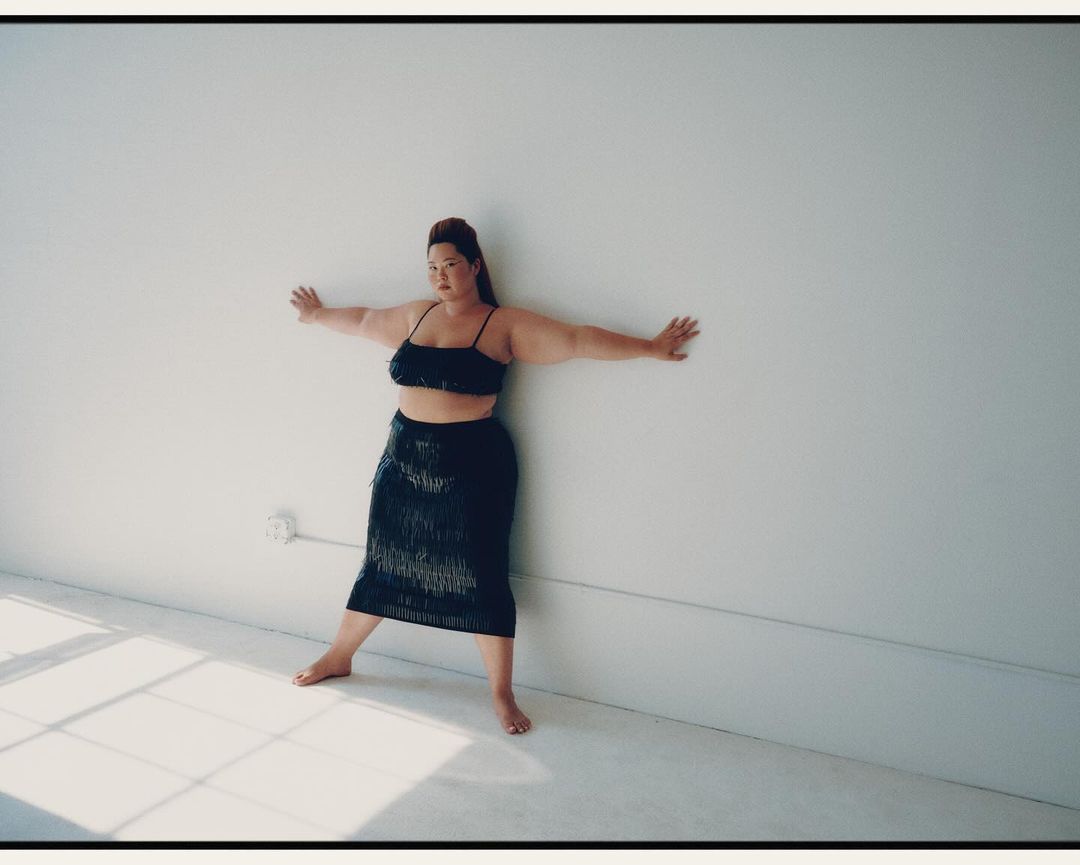 Adult female poses with arms outstretched against a white wall, wearing a black fringe skirt and crop top in a minimalist indoor setting.