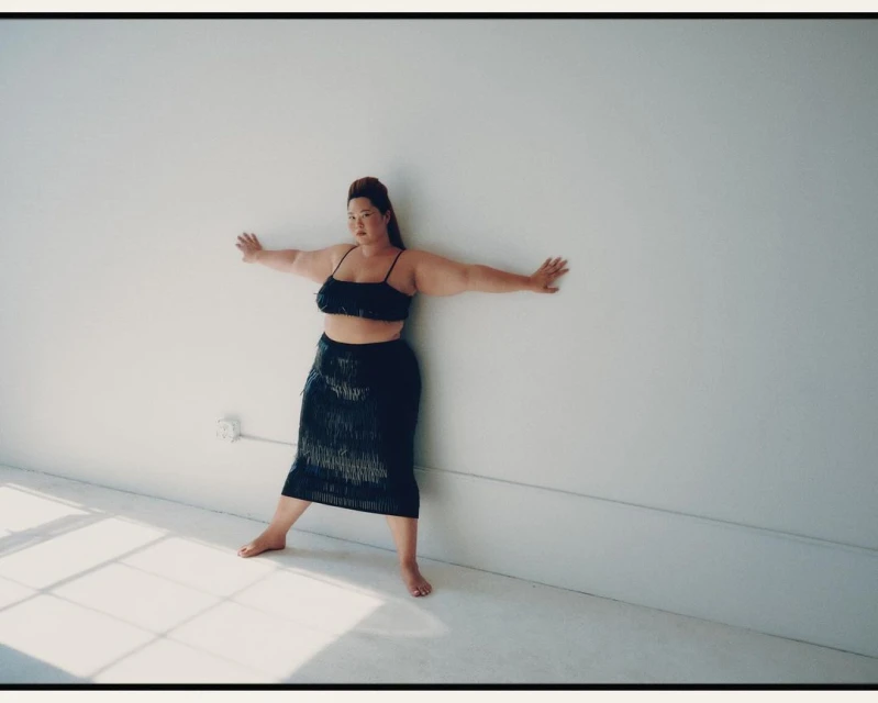 Adult female poses with arms outstretched against a white wall, wearing a black fringe skirt and crop top in a minimalist indoor setting.