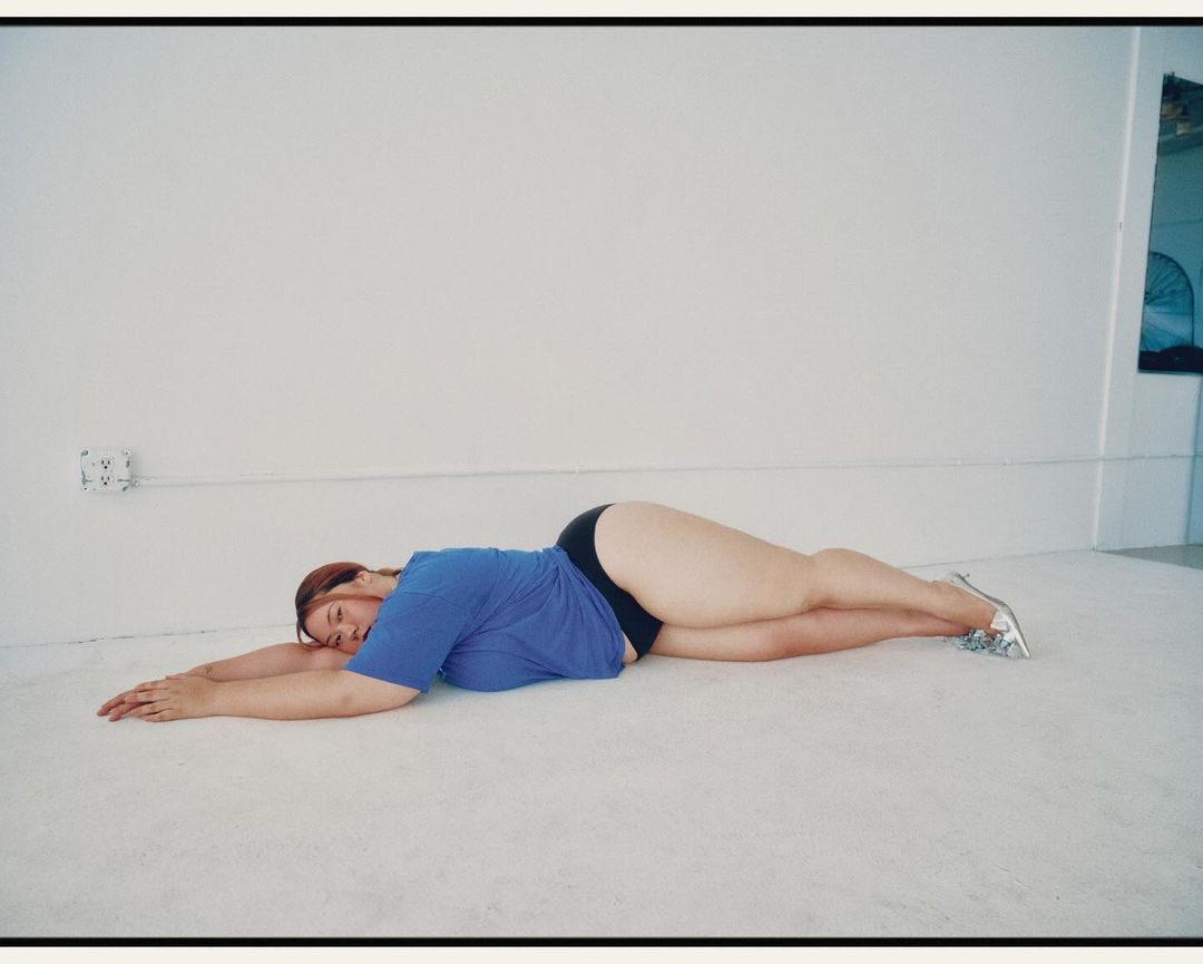 A woman with red hair lies on her side on a white floor wearing a blue shirt and black shorts. She has silver shoes and is indoors.