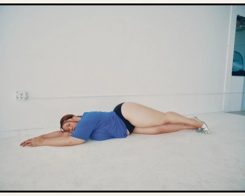 A woman with red hair lies on her side on a white floor wearing a blue shirt and black shorts. She has silver shoes and is indoors.