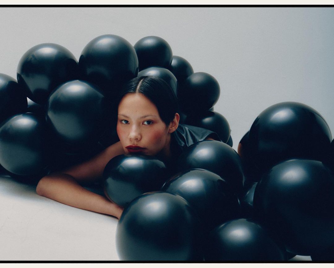Asian woman lies amid a collection of glossy black balloons in a photo studio with simple lighting and dark red lipstick. 