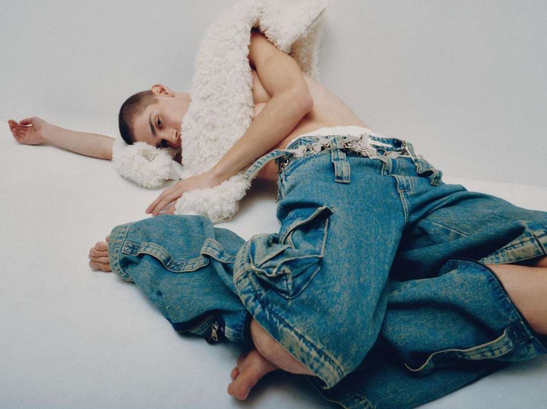 Androgynous person poses in denim overalls and a white, furry stole in a bright studio environment for a fashion editorial.