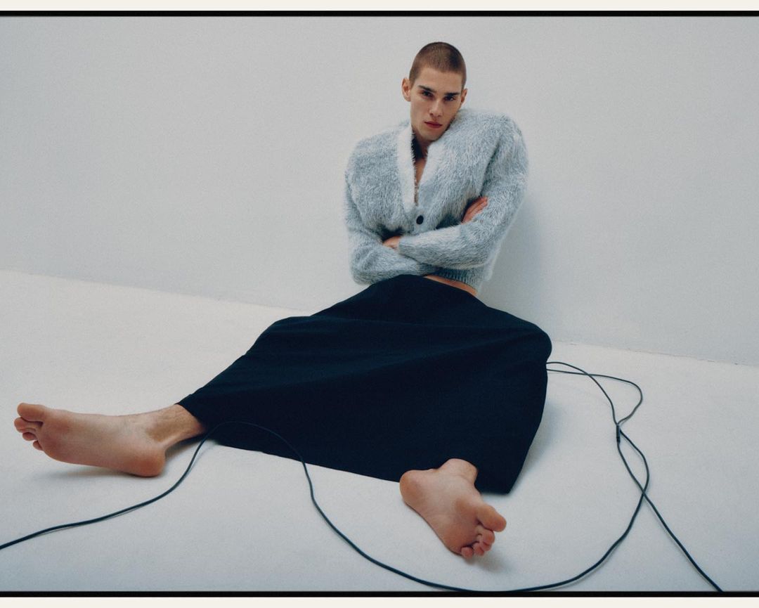 A young adult male with a buzzcut is sitting on the floor wearing a fuzzy grey cardigan and a black skirt with his arms crossed. 